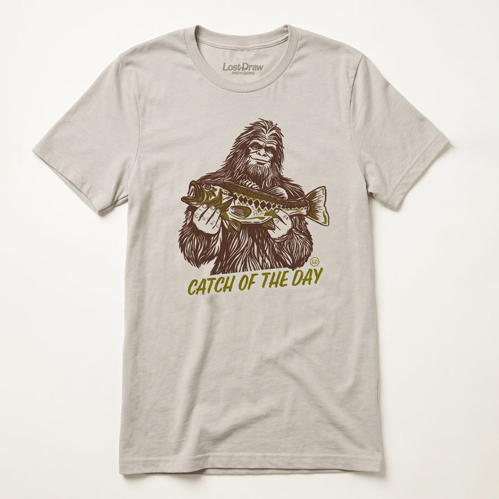 A light beige crewneck t-shirt featuring a graphic of Bigfoot holding a fish, with the text &