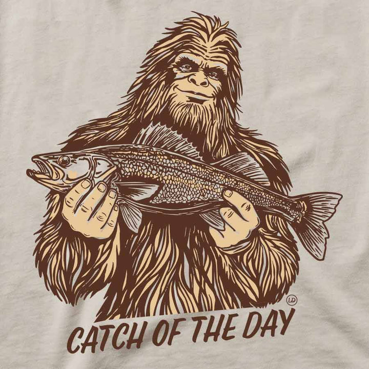 A close-up of a graphic print on a light beige fabric, featuring a brown line art illustration of Bigfoot holding a walleye fish with the text &