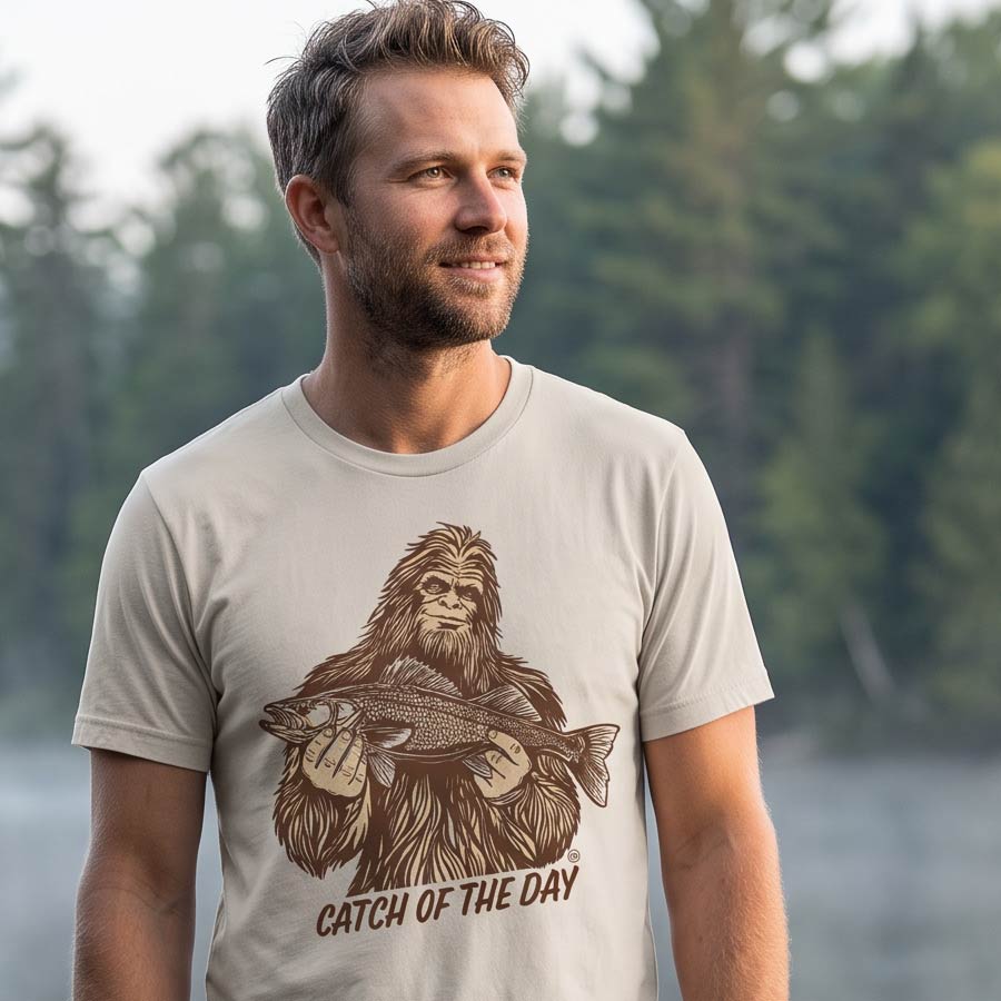 Man wearing the Dust Catch of the Day Walleye Tee on a lakeside dock, with the Bigfoot fishing graphic visible on the front.
