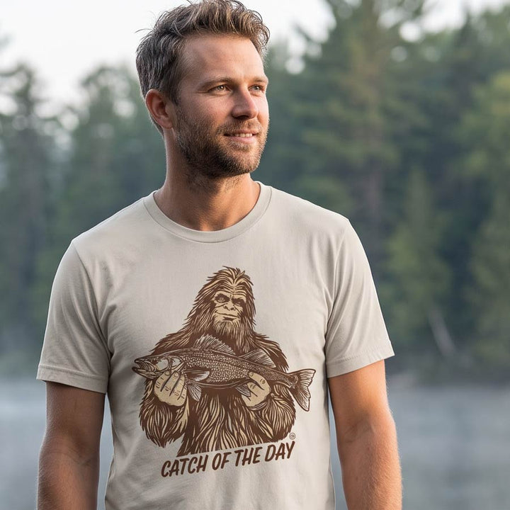 Man wearing the Dust Catch of the Day Walleye Tee on a lakeside dock, with the Bigfoot fishing graphic visible on the front.

