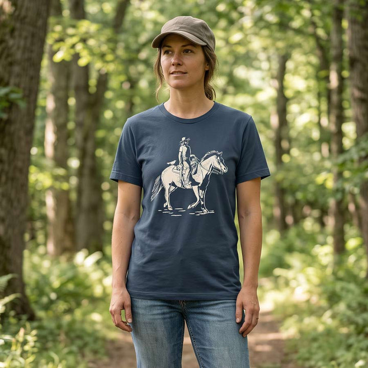 Woman wearing the Vintage Navy Dust &amp; Reins Tee on a wooded trail, showing the cream cowgirl and horse graphic.