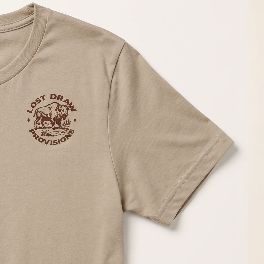 A close-up of a beige short-sleeve t-shirt featuring a brown circular graphic on the left chest with a bison and the text &