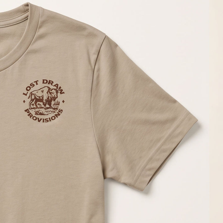 A close-up of a beige short-sleeve t-shirt featuring a brown circular graphic on the left chest with a bison and the text &