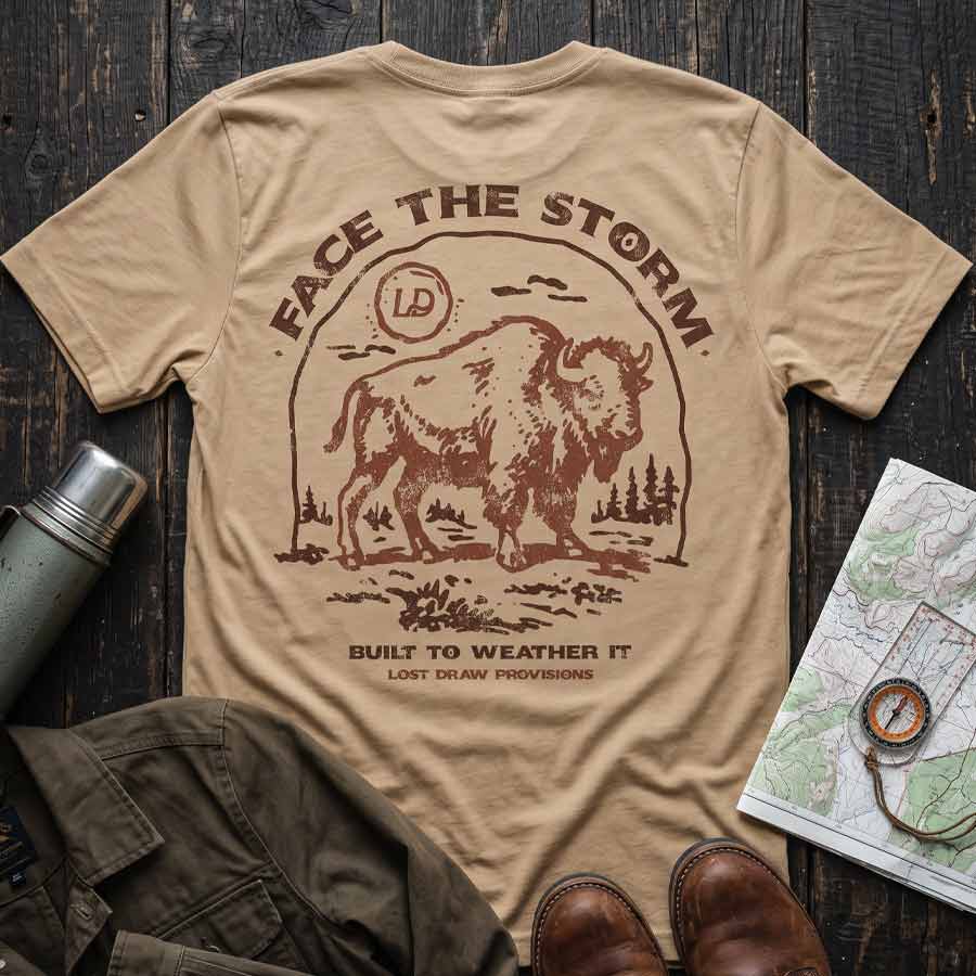 Tan Face the Storm buffalo graphic tee displayed with hiking boots, a canvas jacket, and vintage outdoor accessories.