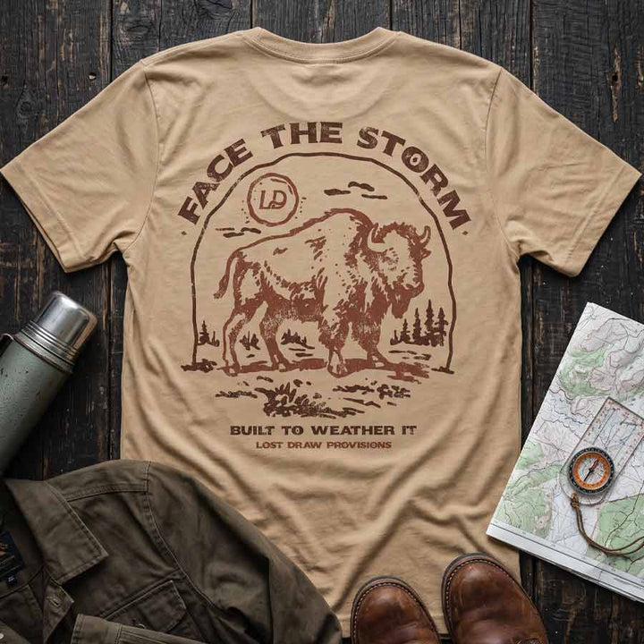 Tan Face the Storm buffalo graphic tee displayed with hiking boots, a canvas jacket, and vintage outdoor accessories.