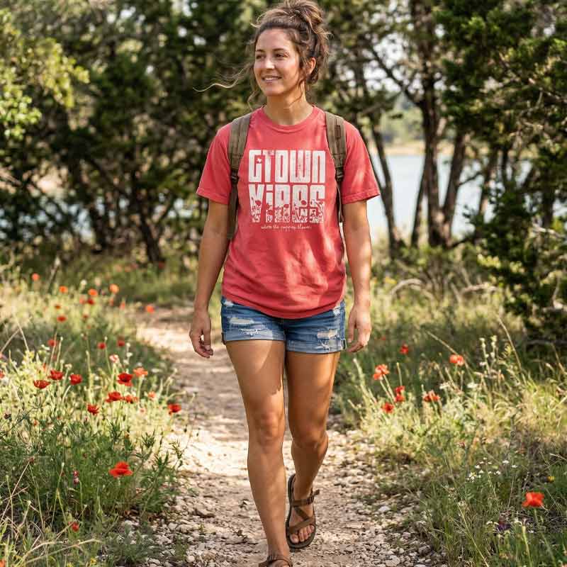 Woman walking on a trail with flowers and trees in the background Woman wearing Georgetown Texas Vibes Tee, printed on a classic Comfort Colors 1717 Paprika t-shirt.