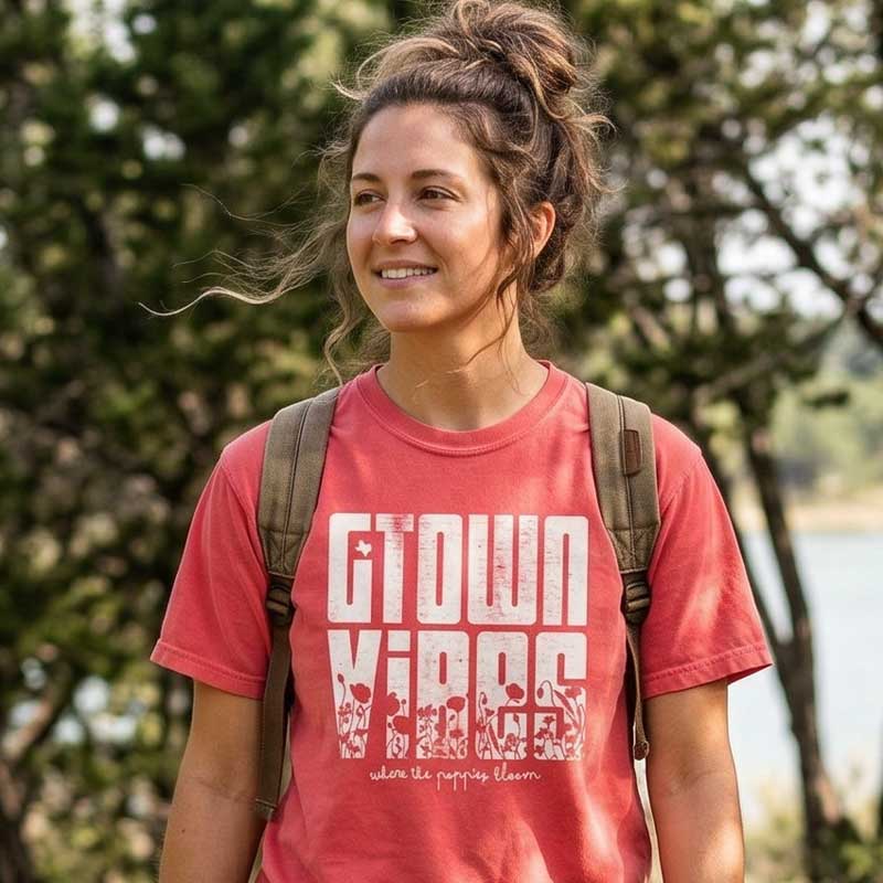 Woman wearing a  Georgetown Texas Vibes  t-shirt with text, standing outdoors with trees and water in the background