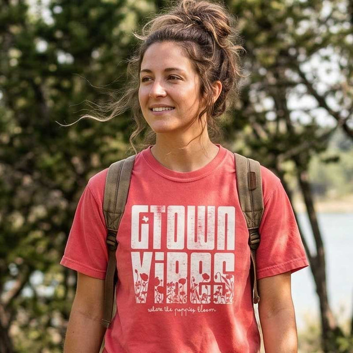 Woman wearing a  Georgetown Texas Vibes  t-shirt with text, standing outdoors with trees and water in the background