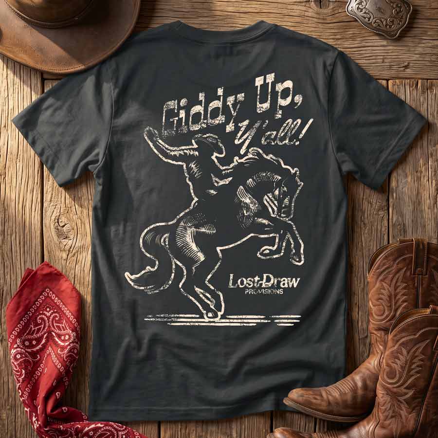 A dark charcoal gray graphic t-shirt laid flat on a rustic wooden surface, surrounded by western-themed props including a cowboy hat, a red bandana, and brown leather cowboy boots. The back of the t-shirt features a distressed white graphic of a cowboy riding a bucking horse, with the text &