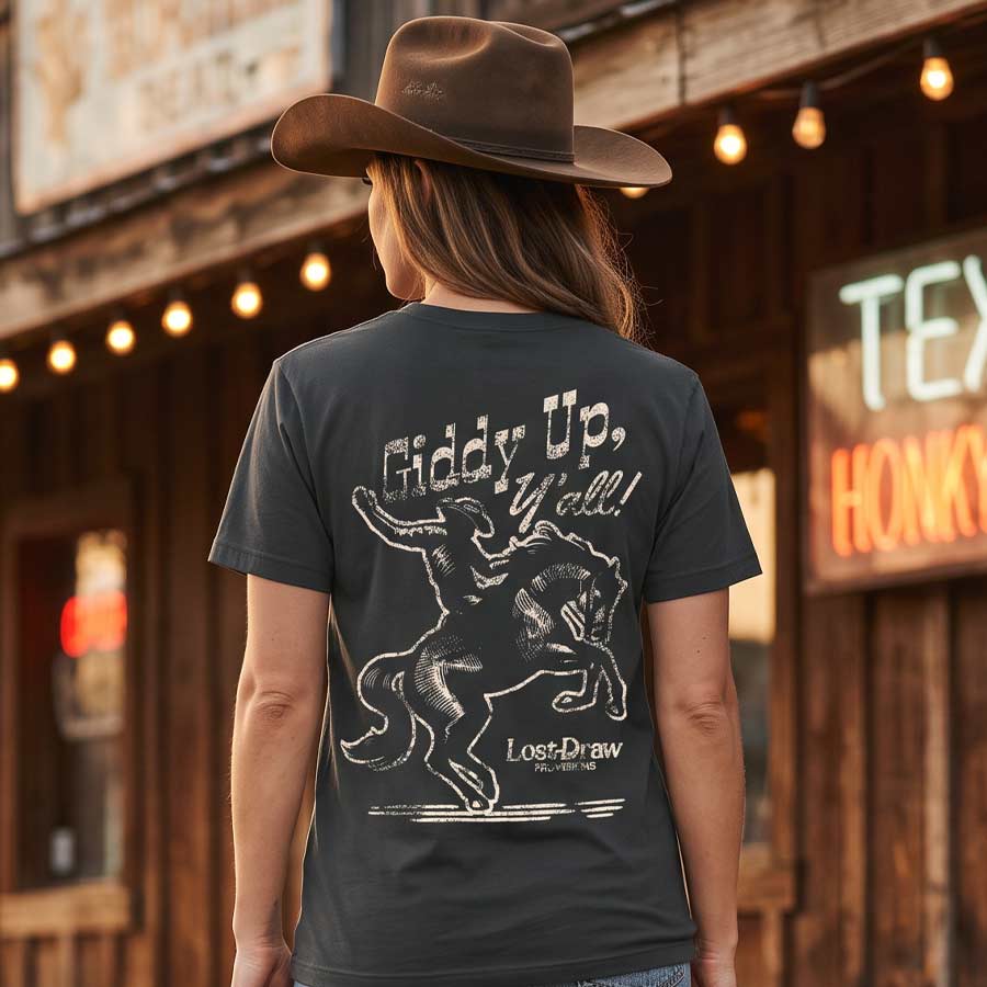A woman wearing a brown cowboy hat and a dark charcoal gray graphic t-shirt is shown from behind, standing in front of a rustic wooden saloon with warm string lights. The t-shirt features a white distressed graphic of a cowboy riding a bucking bronco with the text 'Giddy Up, Yell!' and 'Lost Draw'.