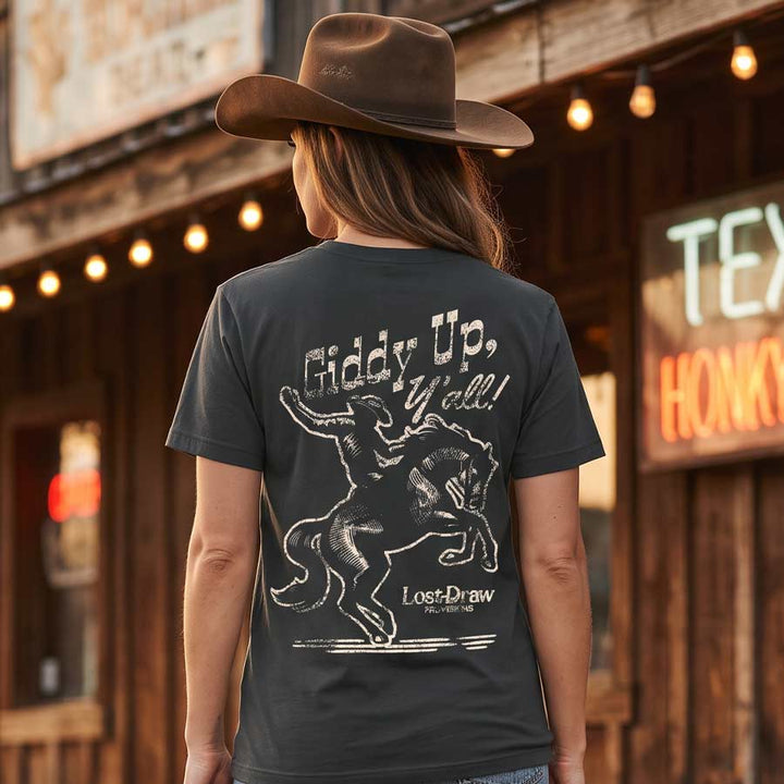 A woman wearing a brown cowboy hat and a dark charcoal gray graphic t-shirt is shown from behind, standing in front of a rustic wooden saloon with warm string lights. The t-shirt features a white distressed graphic of a cowboy riding a bucking bronco with the text &
