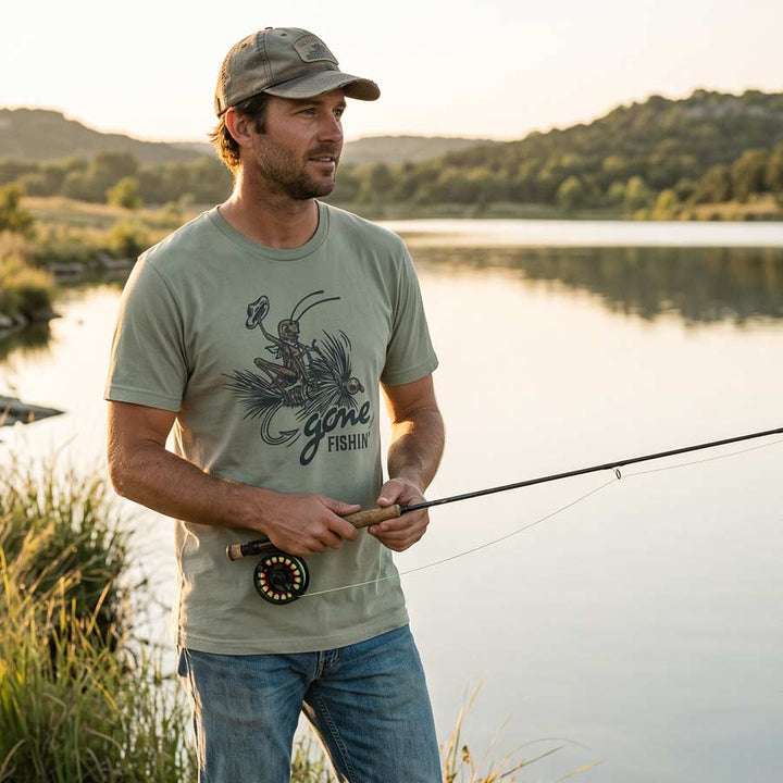 A man stands by a lake at sunset wearing a light sage green t-shirt with a &