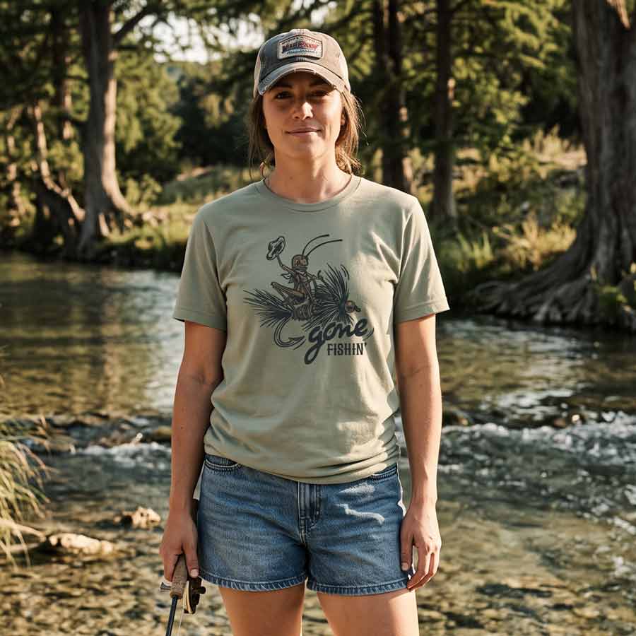 A woman stands in a shallow stream wearing an olive green graphic T-shirt with a fly-fishing design and the words 'Gone Fishing,' paired with denim shorts and a baseball cap, against a natural forest stream background.
