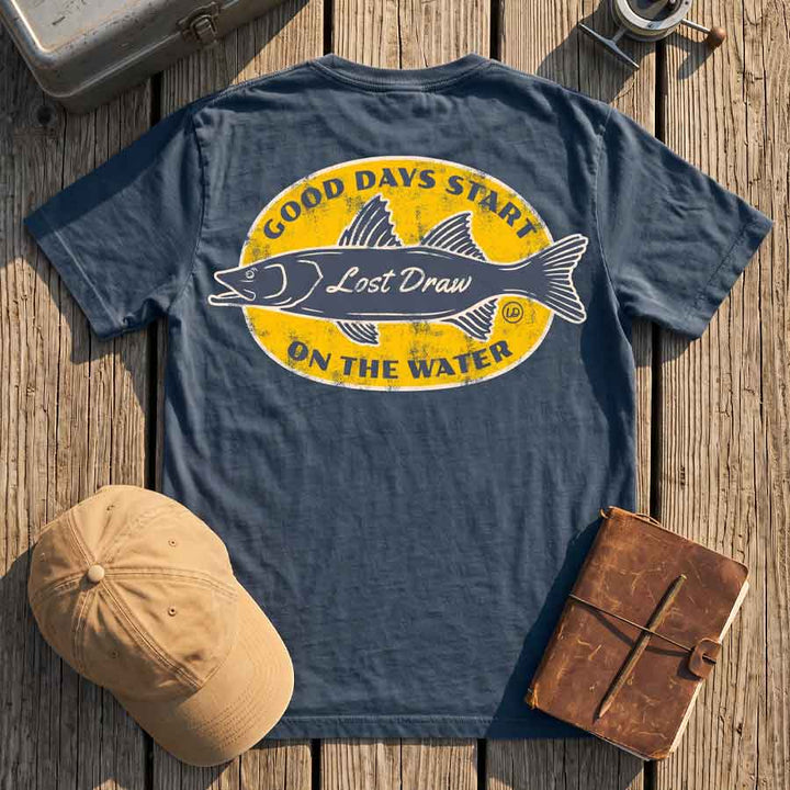 Vintage Navy On the Water fishing tee displayed with a tackle box, fishing reel, and canvas cap on a wooden dock.