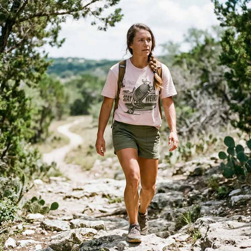 Woman hiking on a trail wearing soft pink Stay Weird 512 boxy t-shirt with armadillo and Austin skyline graphic