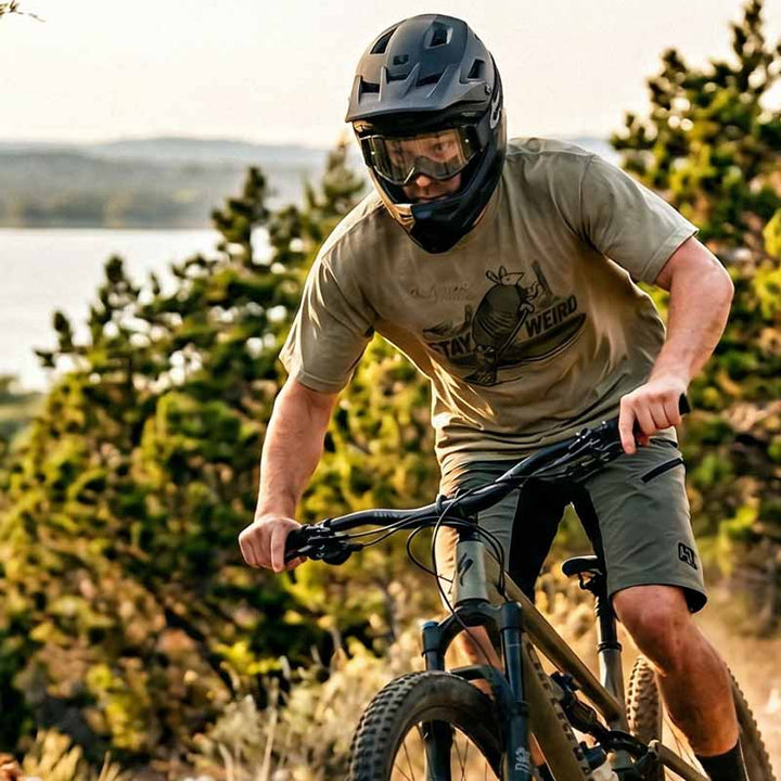 Man riding a mountain bike wearing a khaki 512 Austin armadillo t-shirt, on a trail with trees and a lake in the background