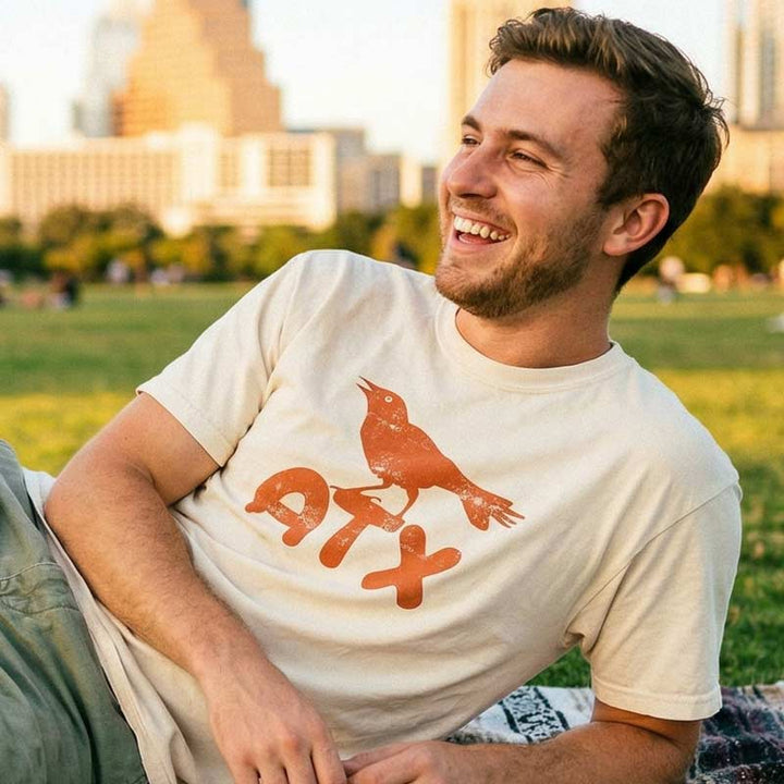 Man wearing a Austin grackle graphic tee with distressed orange print on ivory shirt, sitting outdoors.