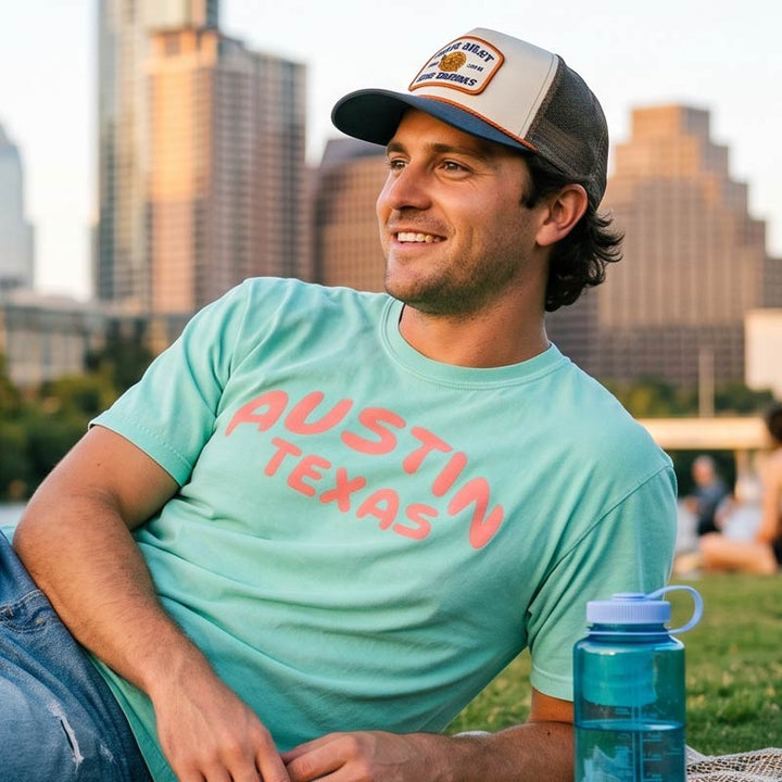 Man wearing a Chalky Mint Comfort Colors 1717 Austin t-shirt with coral graphic at Zilker Park with the Austin skyline in the background.