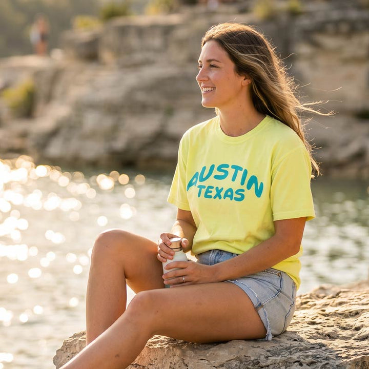 Woman wearing a Neon Lemon Austin graphic t-shirt near Lake Travis at golden hour. Aqua Austin Texas lettering visible on the front of the tee.