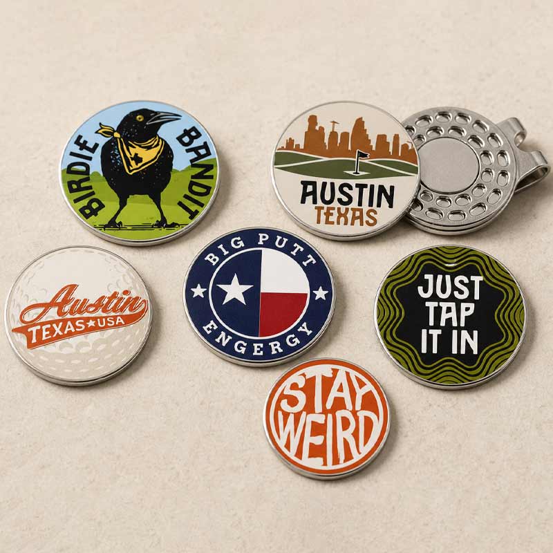 Six round enamel golf tags with humorous and Texas-themed designs, arranged on a light beige textured background. The designs include a bird with a golf club, the Austin skyline with a flag, the Austin Texas USA script, the Texas flag with 'Big Putt Engergy', a green wavy pattern with 'Just Tap It In', and an orange circle with 'Stay Weird'.
