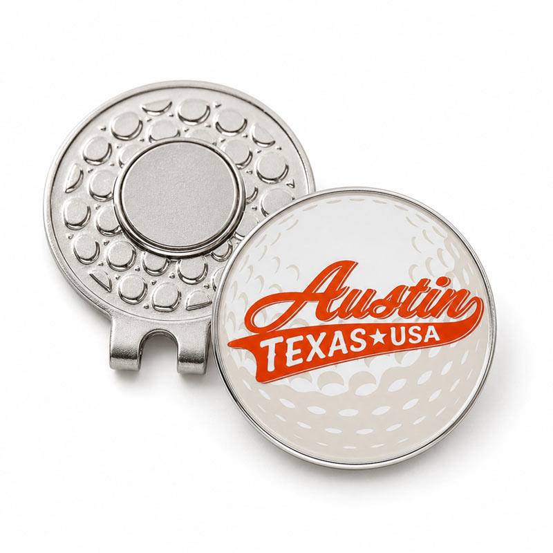 A silver metal golf ball clip shown open, with one side featuring a textured circular design and the other side displaying a golf ball pattern background with the words &