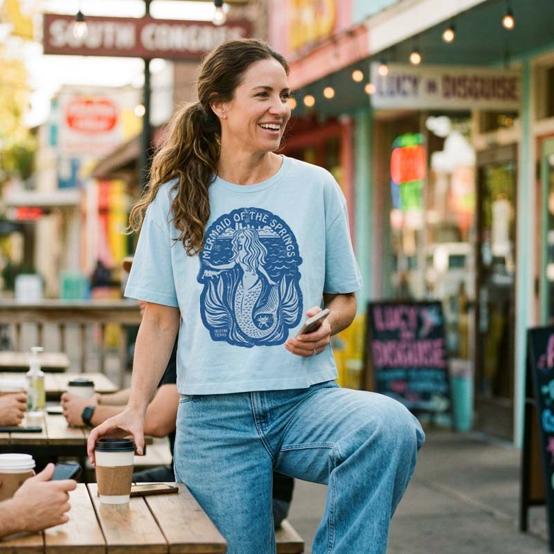 Woman wearing Barton Springs Mermaid women’s boxy t-shirt with mermaid graphic, sitting outdoors in a casual setting.