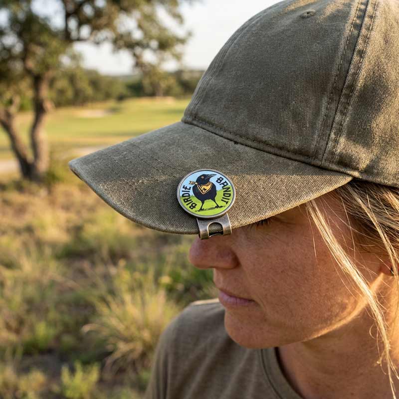 A woman outdoors wearing an olive green cap with a metal clip holding a round black-and-white 'Buckeye Bear' badge against her brow, with a blurred grassy field and trees in the background.