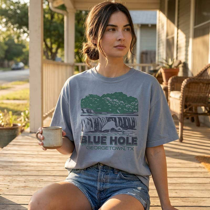 Woman sitting on a wooden deck holding a mug, wearing a boxy Blue Hole Georgetown TX t-shirt in garment-dyed grey with vintage waterfall design and relaxed cropped fit