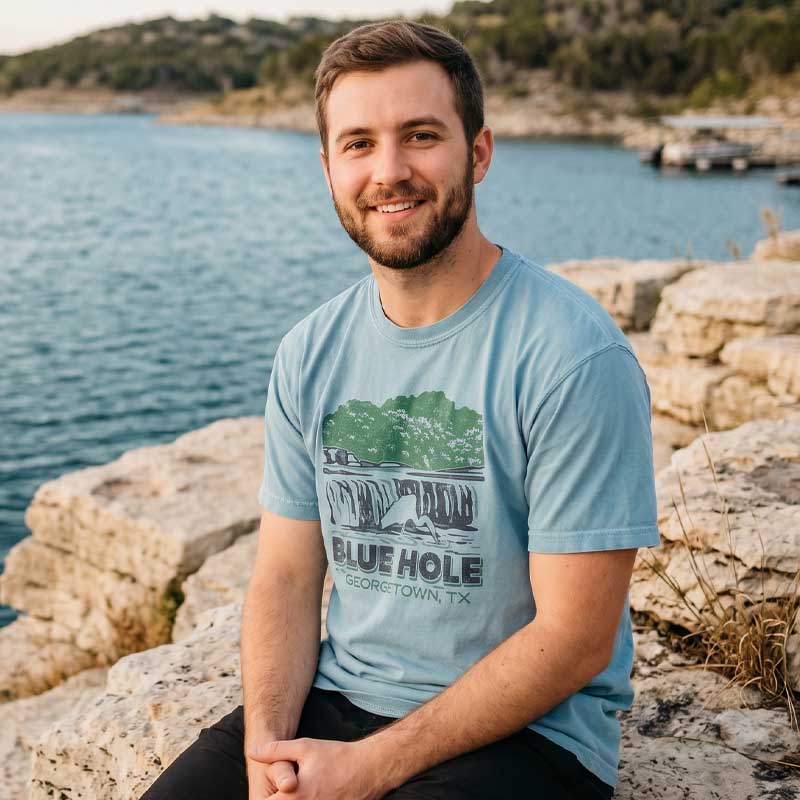 Man wearing a blue t-shirt with 'Blue Hole Georgetown, TX' design by a lake.