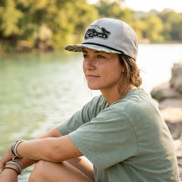 Woman wearing the Carry-On Austin Camo Hat sitting on a river bank.
