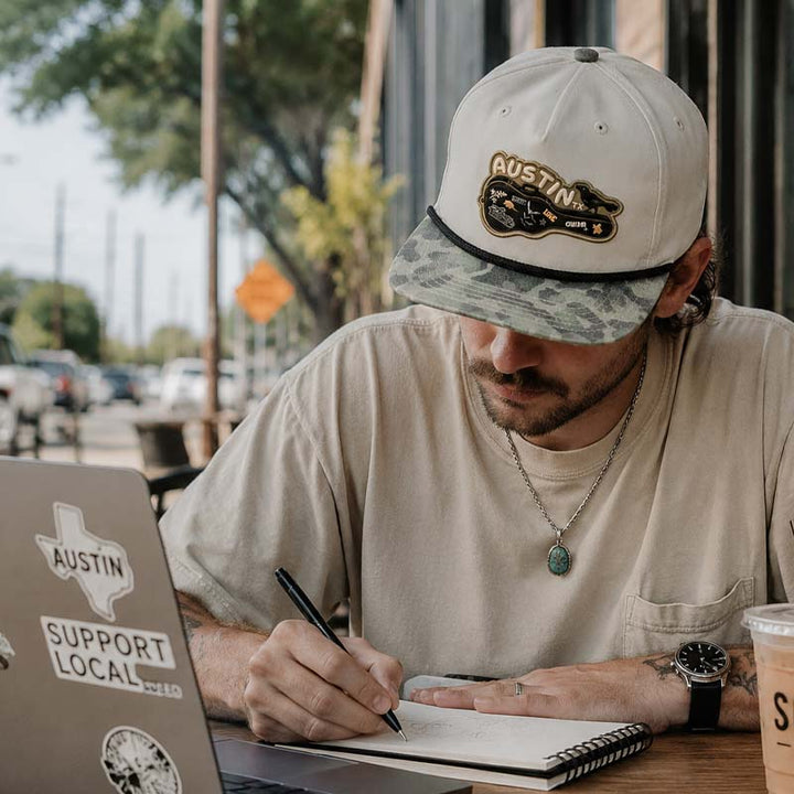 Person wearing the Carry-On Austin Camo Hat at an outdoor Austin coffee shop
