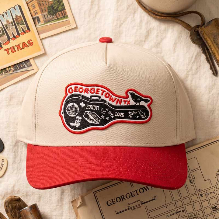 Carry-On Georgetown Hat styled in a flat lay with vintage Texas-inspired travel props, featuring the Georgetown guitar-case patch on a cream and red five-panel cap.
