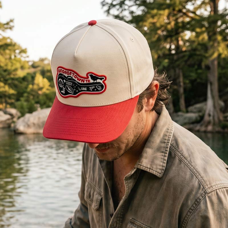 Person wearing the Carry-On Georgetown Hat outdoors near the water, featuring a cream and red Georgetown Texas five-panel cap with guitar-case patch and grackle detail.