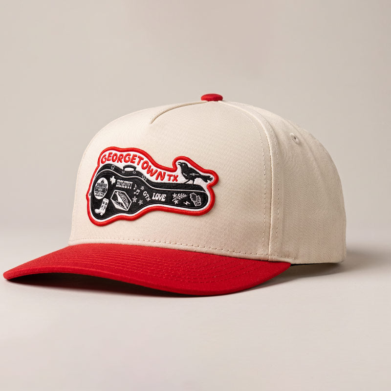 Angled studio photo of the Carry-On Georgetown Hat in cream and red, showcasing the Georgetown guitar-case patch, structured five-panel fit, and curved red bill.
