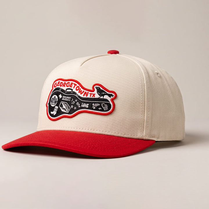 Angled studio photo of the Carry-On Georgetown Hat in cream and red, showcasing the Georgetown guitar-case patch, structured five-panel fit, and curved red bill.
