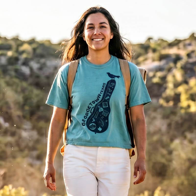 Woman wearing a teal t-shirt with a graphic design, standing outdoors with a blurred natural background