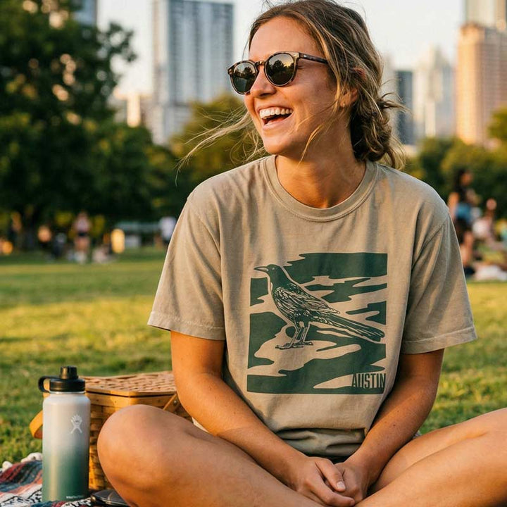 Woman sitting in a park wearing a Sandstone unisex t-shirt with Austin camo style graphic and bird design