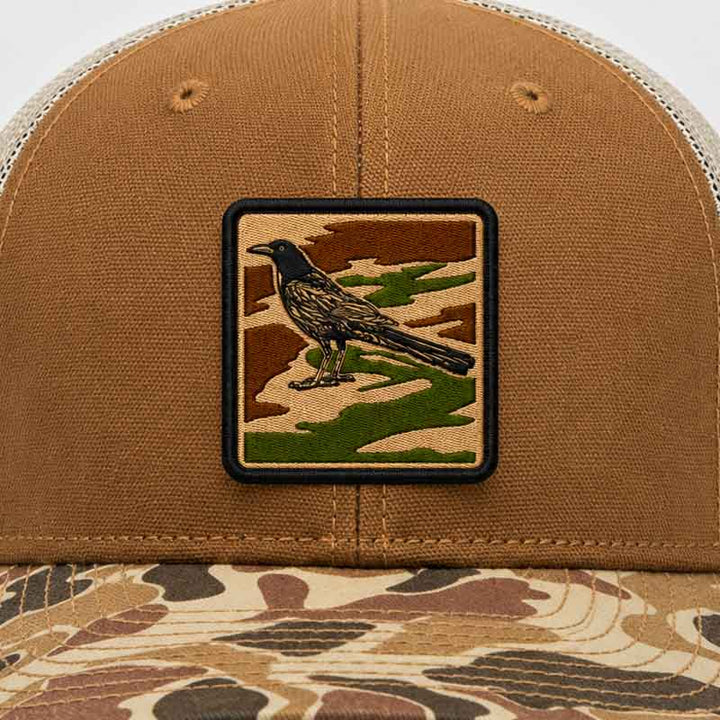 Close up of Saddle and duck camo DRI DUCK trucker hat with UV printed grackle camouflage patch designed to look embroidered