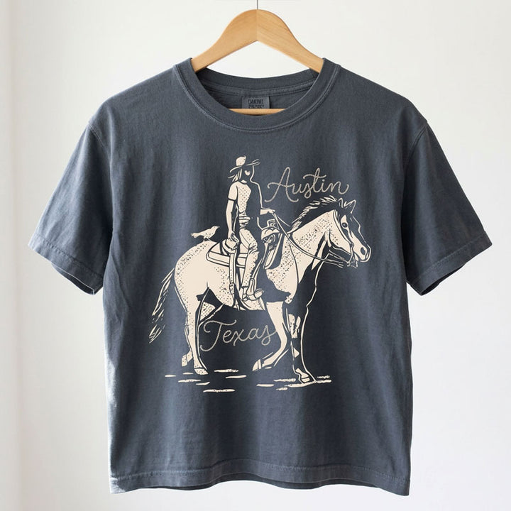 Women’s garment-dyed boxy tee in washed black with western Austin graphic, hanging on a wooden hanger against a white background.