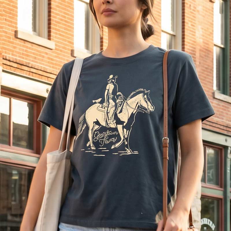 Person wearing a navy blue t-shirt with a graphic design of a person on a horse, standing in front of a brick building.