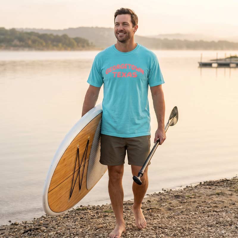 Man holding a paddleboard and paddle on a lakeside with a scenic background wearing Georgetown Texas pop lettering T-shirt on lagoon Comfort Colors 1717 with coral text graphic