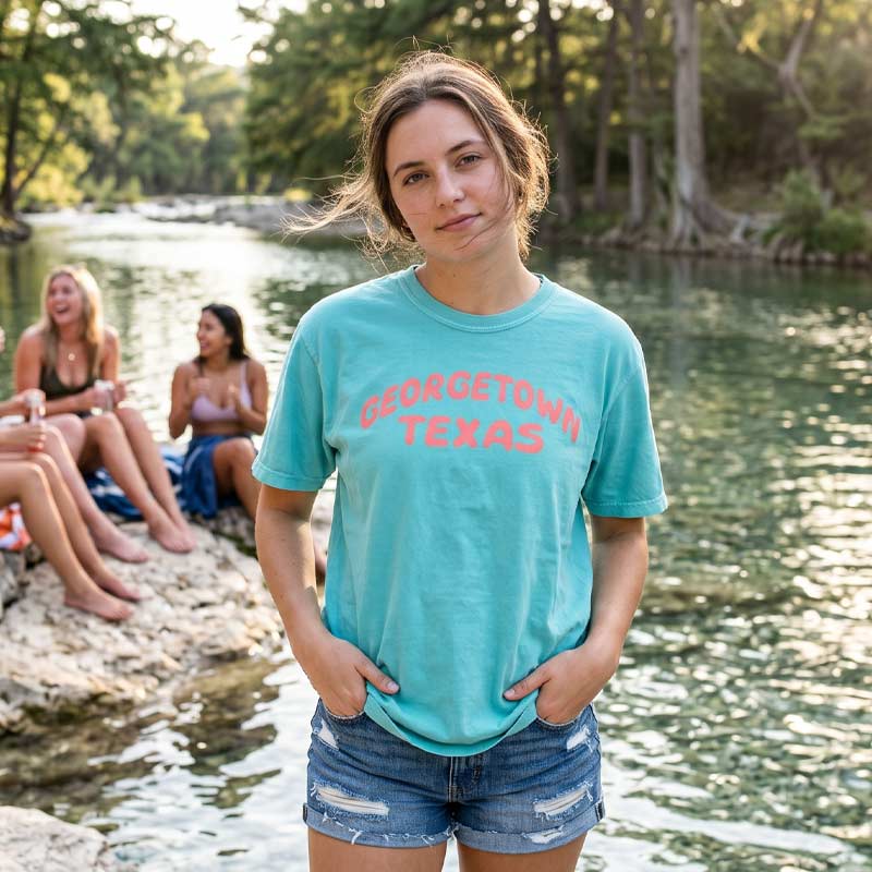 Woman wearing Georgetown Texas pop lettering T-shirt on lagoon Comfort Colors 1717 with coral text graphic by a river with friends in the background.