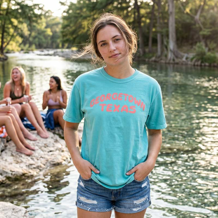 Woman wearing Georgetown Texas pop lettering T-shirt on lagoon Comfort Colors 1717 with coral text graphic by a river with friends in the background.