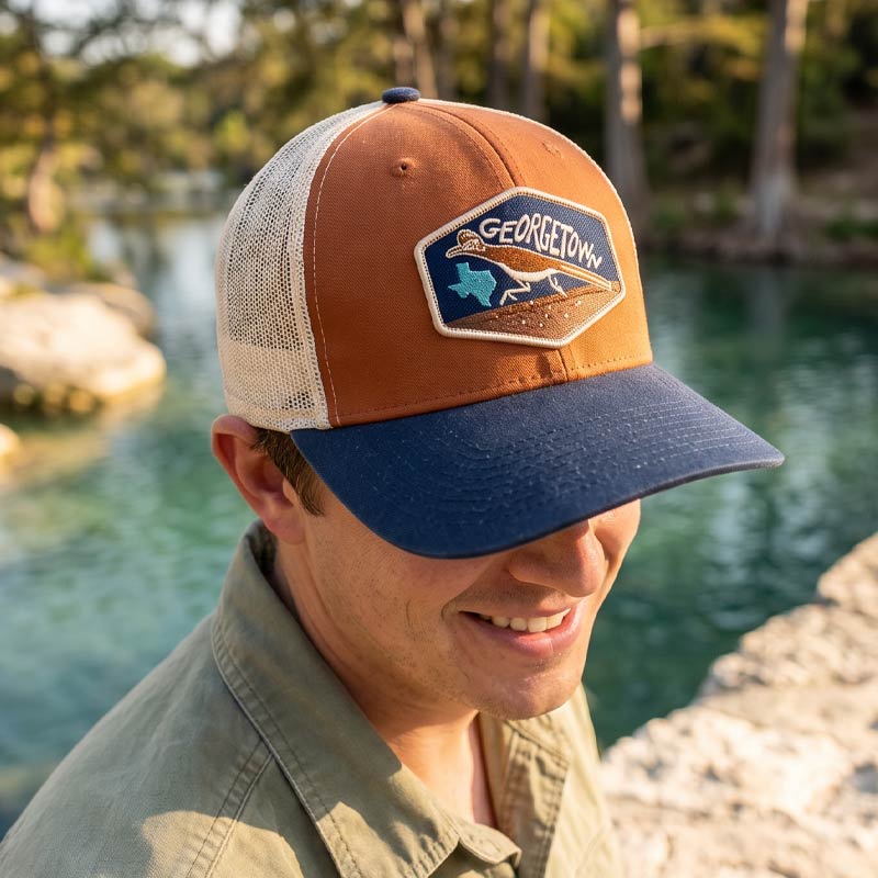 Man wearing the Georgetown Road Runner trucker hat by a body of water with trees in the background