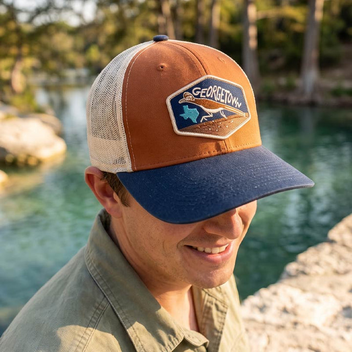 Man wearing the Georgetown Road Runner trucker hat by a body of water with trees in the background