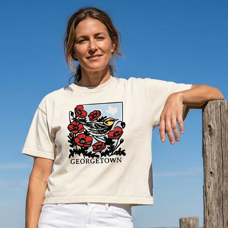 Woman wearing Golden-cheeked warbler graphic tee with Texas outline by Gusto Graphic Tees, standing against a clear blue sky.