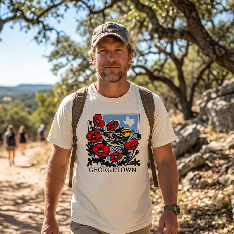 Man wearing Georgetown Warbler ivory t-shirt, standing outdoors with trees and rocks in the background.