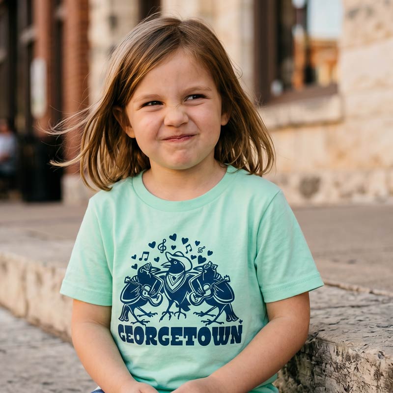 Close-up of girl wearing Georgetown Grackle Gang youth t-shirt in mint featuring navy grackle band artwork in Georgetown, Texas.
