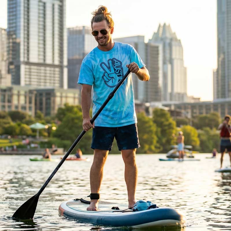Man on a paddleboard in a city park with tall buildings in the background wearing Groovy Austin Blue Lagoon tie-dye graphic tee
