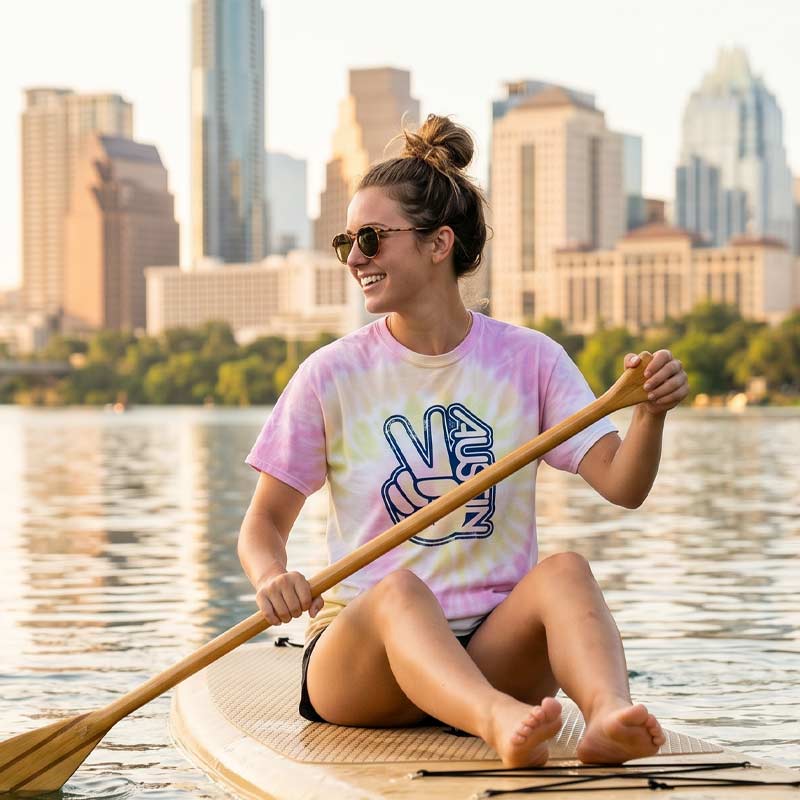 Woman on a paddleboard with a city skyline in the background wearing the Groovy Austin Desert Rose tie-dye t-shirt featuring a retro Austin peace sign graphic during a sunny day in Austin, Texas.